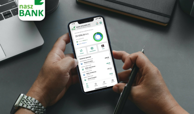 Zdjęcie przedstawia dłonie osoby trzymającej smartfon z uruchomioną aplikacją mobilną Nasz Bank. Urządzenie znajduje się na ciemnoszarym biurku, w otoczeniu przedmiotów biurowych.