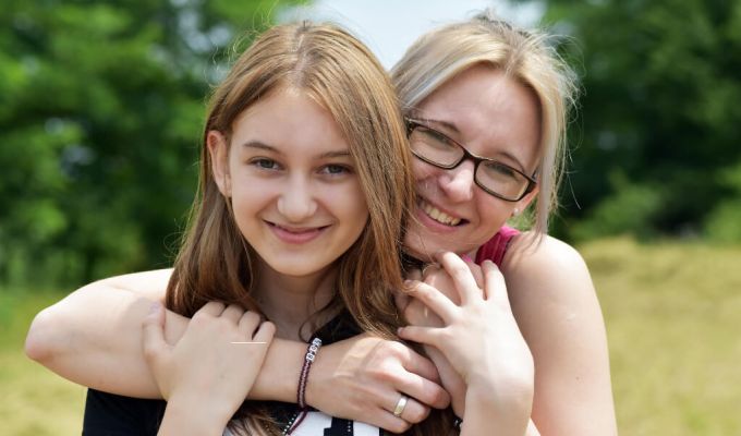Uśmiechnięta kobieta w okularach obejmuje młoda dziewczynę. Obie stoją na tle zieleni.