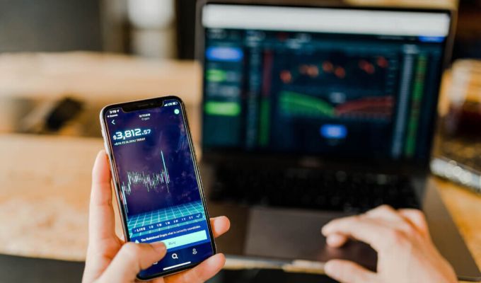 widoczne dwie dłonie, w lewej telefon z otwartą aplikacją finansową, druga dłoń na klawiaturze laptopa.
