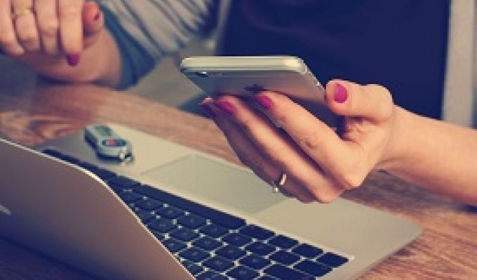 Kobieta siedząca przy biurku, na którym stoi laptop. W lewej dłoni trzyma telefon.