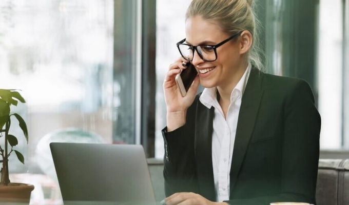 Uśmiechnięta kobieta w okularach i biznesowym stroju rozmawia przez telefon, jednocześnie pracując na laptopie w biurze.