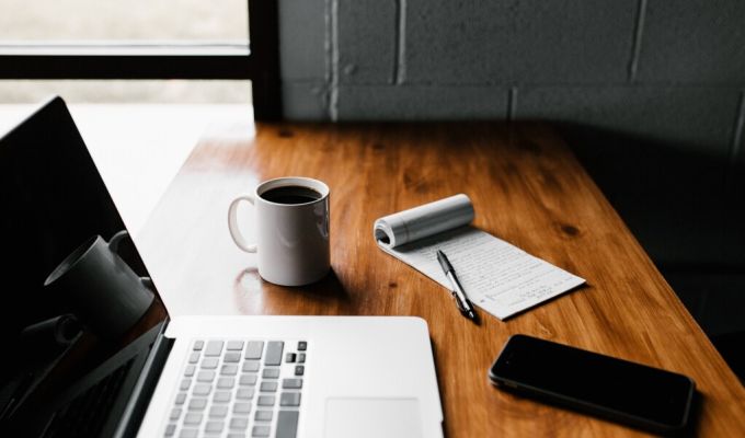 Na zdjęciu jest drewniane biurko na którym jest laptop, telefon i notes