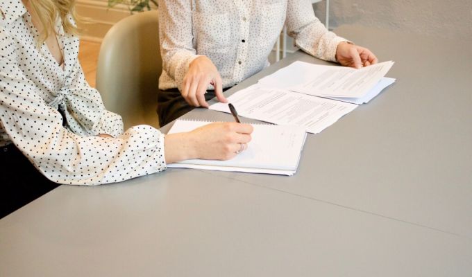 Dwie osoby w strojach biznesowych siedzą przy biurku; jedna z nich pisze w notatniku, a druga przegląda dokumenty papierowe.